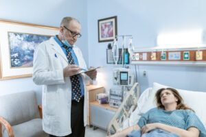 Doctor in white coat checking patient chart in hospital room with medical equipment.