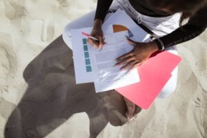 A person reviews and signs documents while sitting on a sandy beach, capturing a relaxed yet focused moment.