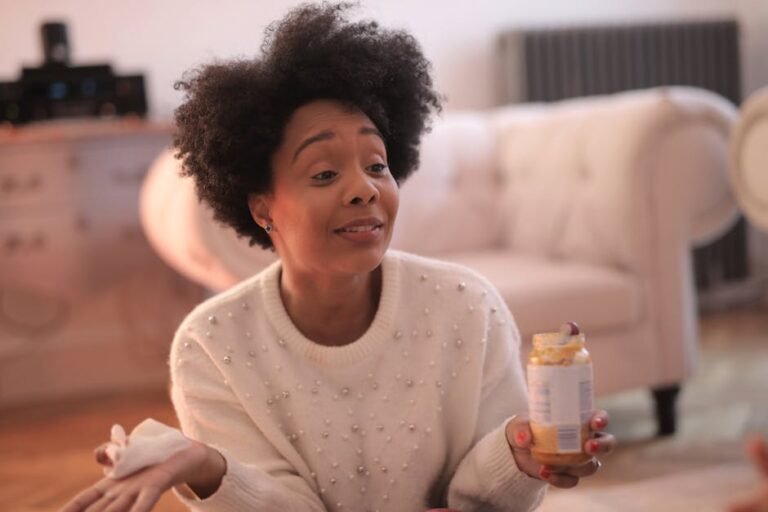 Cheerful African American female in warm casual sweater sitting on floor with jar of baby food in cozy living room and looking away