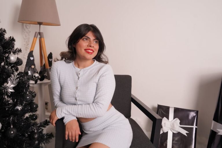 Woman sitting beside a decorated Christmas tree indoors, smiling and enjoying the festive ambiance.