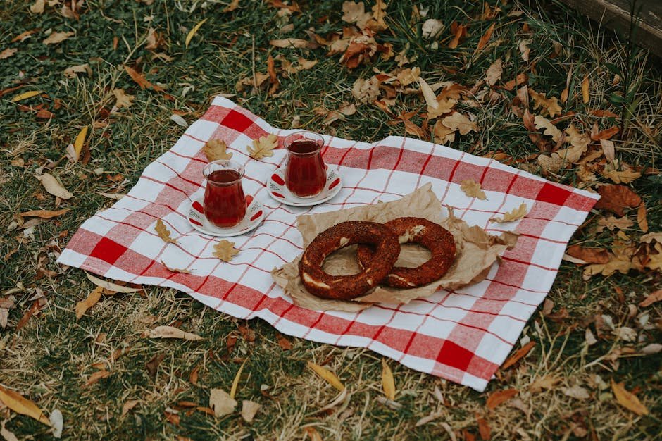 Enjoy a cozy autumn picnic with two cups of tea and simit on a red-checkered cloth on a grassy field.