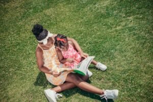 Two young sisters enjoy reading together on a sunny day outdoors in a park, showcasing sibling bonding and leisure.